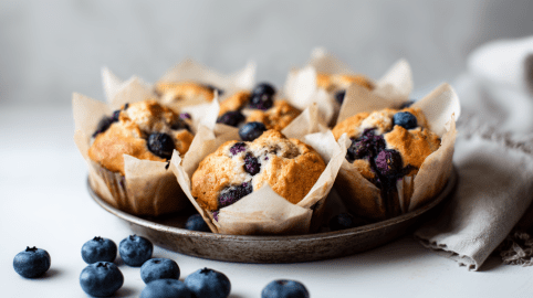 Mehrere Blaubeer-Kardamom-Muffins in einer kleinen Metallschale. Davor liegen einige Blaubeeren.