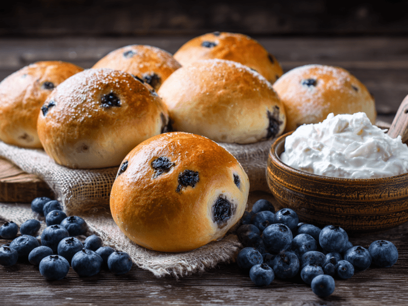 Ein Brett mit sieben Heidelbeer-Quarkbrötchen. Quark und frische Heidelbeeren liegen als Deko außen herum.