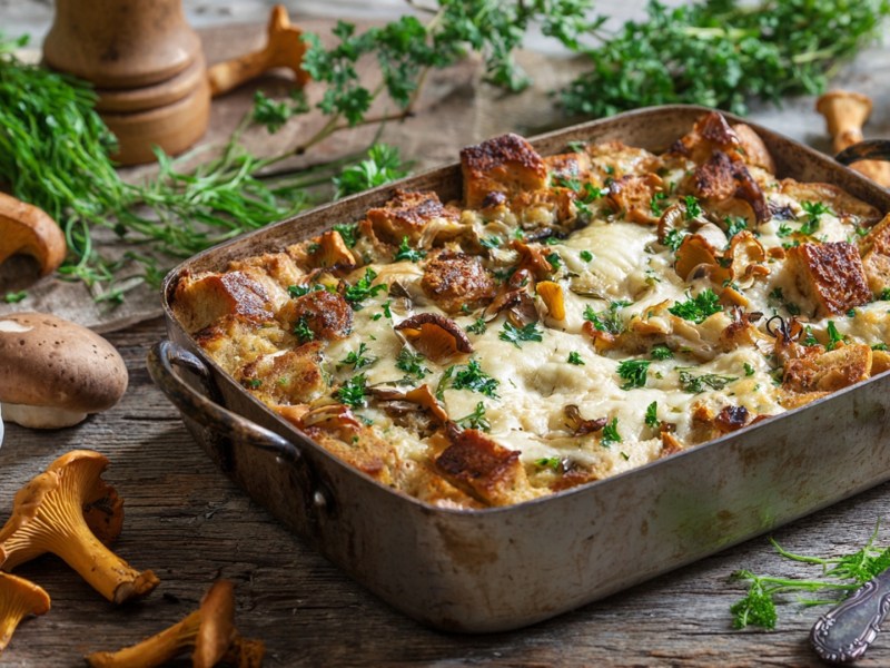 Eine rustikale Ofenform aus Metall, die einen Brotauflauf mit Pfifferlingen enthält und mit frischer gehackter Petersilie garniert ist. Um die Auflaufform herum liegen frische Pfifferlinge, eine Knoblauchknolle und frische Kräuter verteilt.
