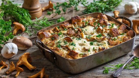 Eine rustikale Ofenform aus Metall, die einen Brotauflauf mit Pfifferlingen enthält und mit frischer gehackter Petersilie garniert ist. Um die Auflaufform herum liegen frische Pfifferlinge, eine Knoblauchknolle und frische Kräuter verteilt.