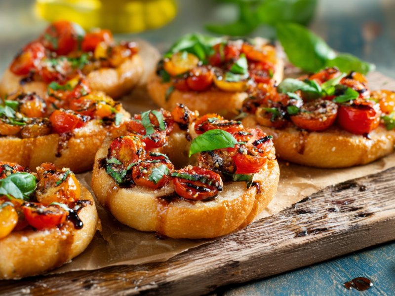 Mehrere Bruschetta-Bagel liegen auf einem Holzbrett. Sie sind mit Tomaten, Basilikum und Balsamico-Essig garniert. Links im Bild sieht man einige Tomaten, rechts liegen noch frische Basilikumblätter.