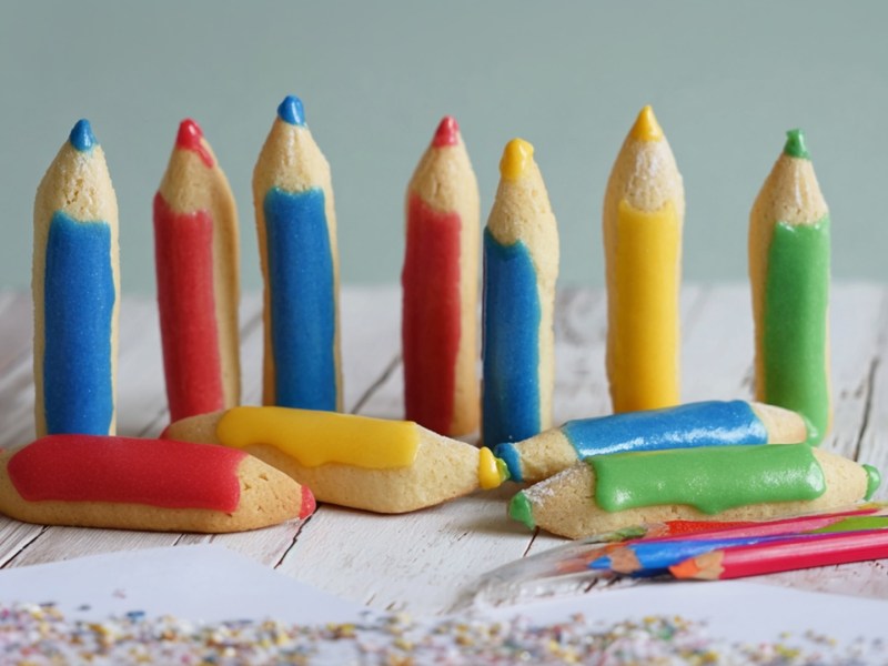 Ein weißer Holztisch, auf dem einige Buntstiftkekse stehen und liegen. Außerdem kann man ein Blatt Papier liegen sehen, auf dem Krümel verteilt sind sowie einige richtige Buntstifte.