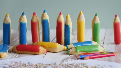 Ein weißer Holztisch, auf dem einige Buntstiftkekse stehen und liegen. Außerdem kann man ein Blatt Papier liegen sehen, auf dem Krümel verteilt sind sowie einige richtige Buntstifte.