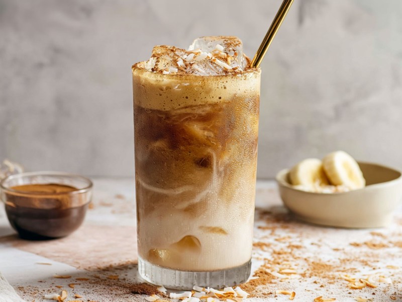 Ein großes Glas mit einem Caramelized Banana Latte. Ein goldener Strohhalm guck aus dem Glas. Im Hintergrund kann man eine Schale mit Bananenscheiben und eine mit Kaffee sehen. Außerdem liegt eine Kokosnuss im Bild. Kokosflocken und Zimt liegen verstreut herum.