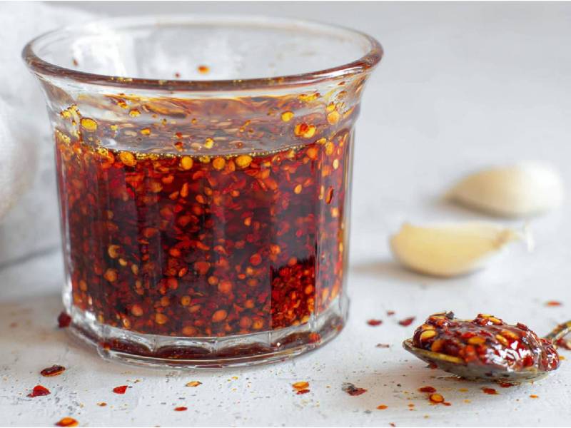 Glas mit Crispy Chili-Öl, Knoblauchzehen im Hintergrund