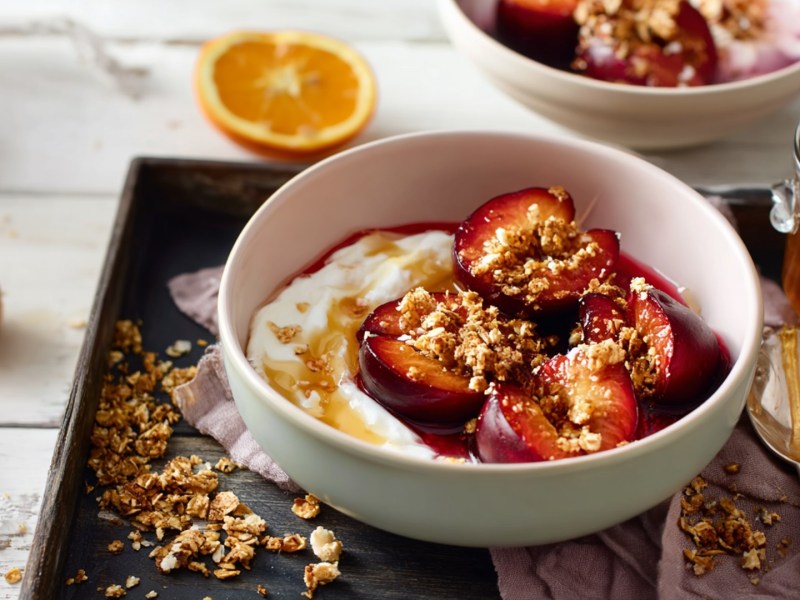 Eine Schüssel mit gebackenen Pflaumen mit Jogurt und Granola. Die Schüssel steht auf einem tiefen Servierblech auf einem hellen Holztisch. Ringsum liegt Granola verstreut, man kann eine halbe Orange, ein Glas Honig, einen Löffel und einen Honiglöffel erkennen.