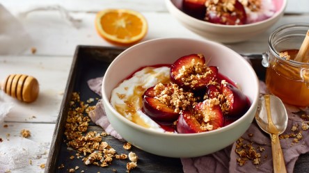Eine Schüssel mit gebackenen Pflaumen mit Jogurt und Granola. Die Schüssel steht auf einem tiefen Servierblech auf einem hellen Holztisch. Ringsum liegt Granola verstreut, man kann eine halbe Orange, ein Glas Honig, einen Löffel und einen Honiglöffel erkennen.