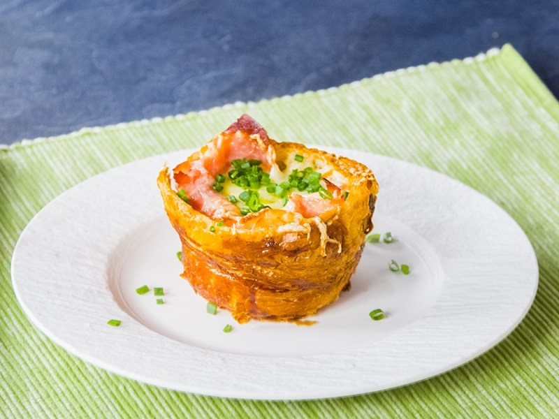Ein gefüllter Croissant-Muffin steht auf einem weißen Teller und ist mit Schnittlauch bestreut. Der Teller steht auf einer grünen Unterlage.