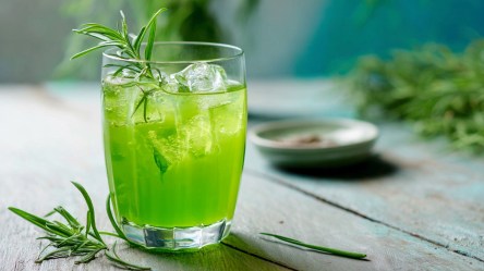 Ein Glas georgische Estragon-Limonade, die auf einer Holzoberfläche steht. Ringsum und im Hintergrund kann frischen Estragon rumliegen sehen.