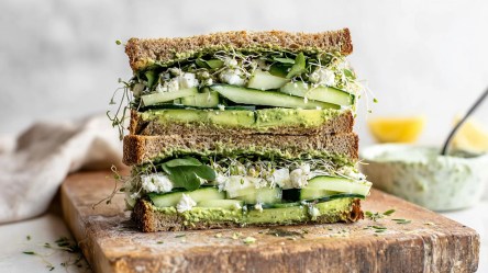 Zwei Hälften eines Green Goddess Sandwich, die aufeinander gestapelt sind. Man sieht sehr gut den Belag aus Avocado, Gurke, Kresse, Feta. Das Sandwich liegt auf einem Holzbrett.