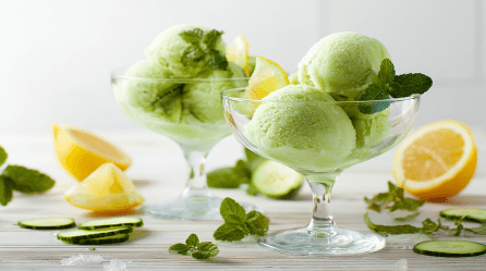 Zwei Dessertgläser mit Gurken-Minz-Sorbet. Um die Gläser verteilt liegen einige Gurkenscheiben, Zitronenstücke und Minzblätter.