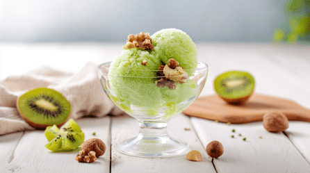 Ein Glas Zitronensorbet auf einem weißen Holztisch. Um das Glas herum liegen einige Nüsse verteilt. Im Hintergrund liegen zwei Kiwihälften.