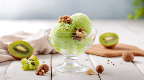 Ein Glas Zitronensorbet auf einem weißen Holztisch. Um das Glas herum liegen einige Nüsse verteilt. Im Hintergrund liegen zwei Kiwihälften.