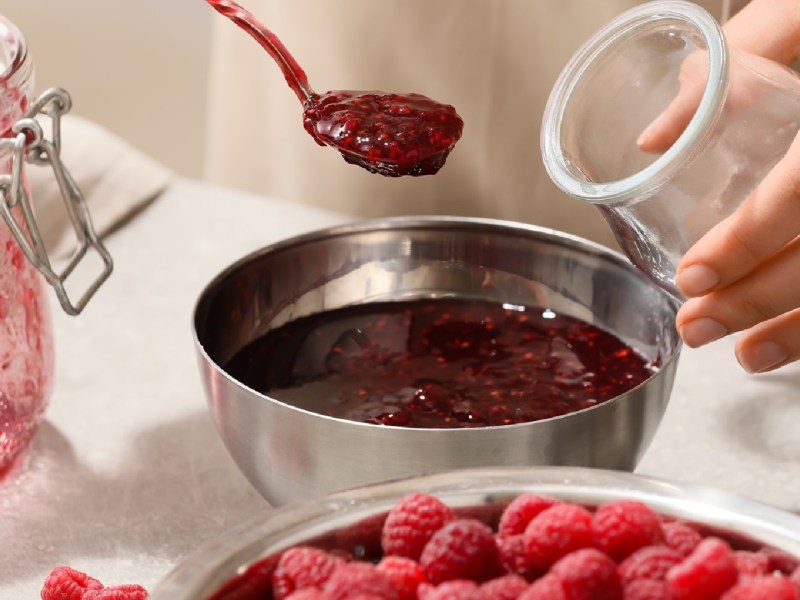 Marmeladen-Zubehör: Ein Mensch füllt Himbeermarmelade aus einer silbernen Schale in ein Glas. Im Vordergrund steht eine Schale mit ganzen, frischen Himbeeren.