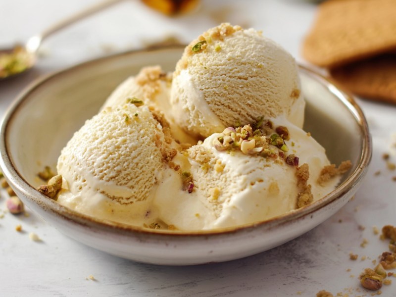 Eine Schüssel mit einigen Kugeln Marzipan-Eis, das mit Amarettini-Bröseln und gehackten Pistazien garniert ist. Im Hintergrund kann man einige Karamellkekse oder Eiswaffeln erkennen sowie einen Löffel mit einigen gehackten Pistazien sehen. Es liegen Keksbrösel und gehackte Nüsse rund um die Schüssel verteilt.