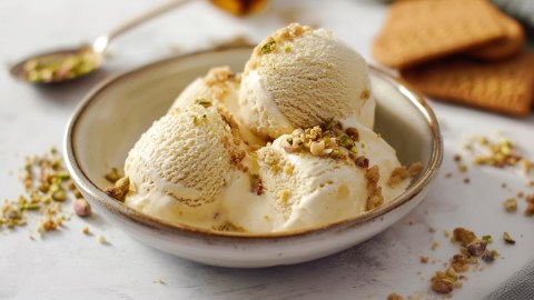 Eine Schüssel mit einigen Kugeln Marzipan-Eis, das mit Amarettini-Bröseln und gehackten Pistazien garniert ist. Im Hintergrund kann man einige Karamellkekse oder Eiswaffeln erkennen sowie einen Löffel mit einigen gehackten Pistazien sehen. Es liegen Keksbrösel und gehackte Nüsse rund um die Schüssel verteilt.