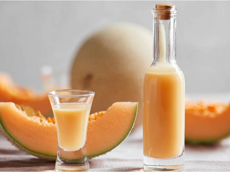 Eine Flasche und ein Glas mit Meloncello, dahinter eine aufgeschnittene Melone