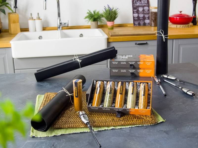 Ein Gewinnspiel-Grill-Set, das schön präsentiert wird. Es liegt eine eingerollte Grillmatte, daneben ein Fleischthermometer und eine geöffnete Schachtel mit einer 5er-Gewürmischung auf einem Tischuntersetzer. Im Vordergrund hängt verschwommen eine grüne Pflanze im Bild.