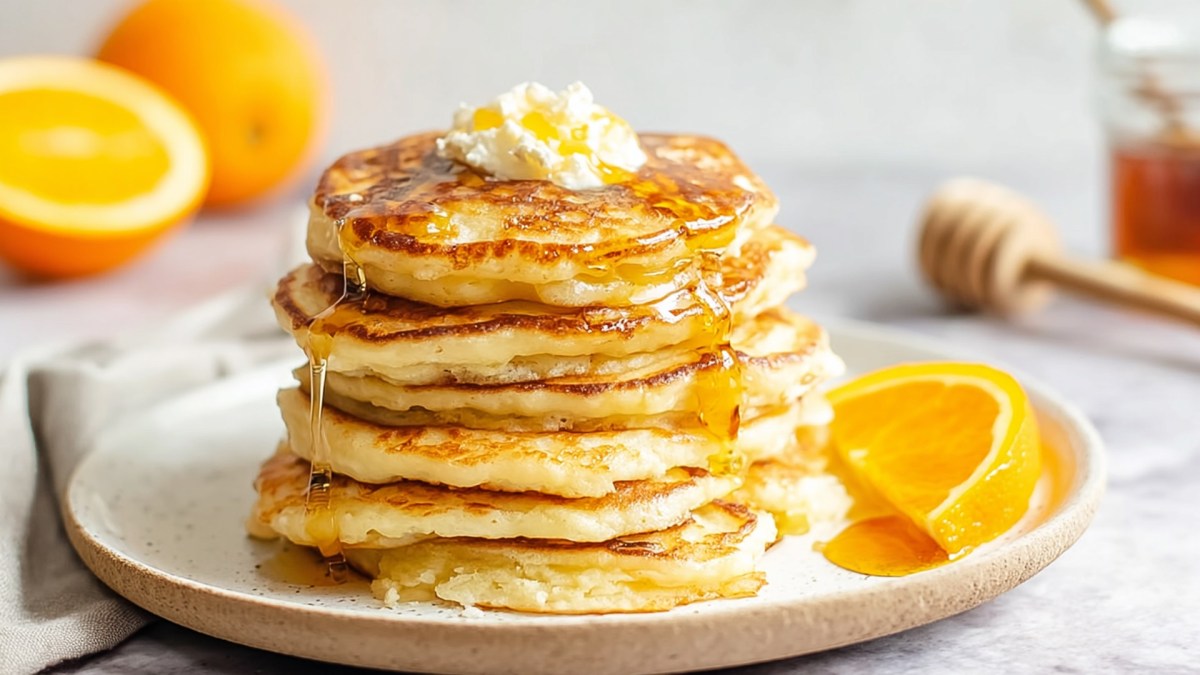 Ein Stapel mit Orangen-Ricotta-Pancakes, die mit einem kleinen Klecks Quark und Honig garniert sind. Neben den Pancakes ist eine halbe Orangenscheibe platziert. Im Hintergrund kann man eine halbe und eine ganze Orange sowie ein Honigglas und einen Honiglöffel erkennen.
