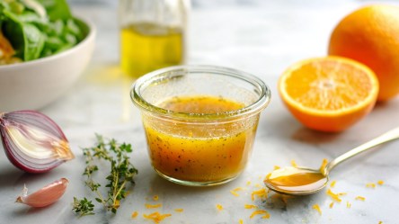 Eine kleine Glasschale mit Orangen-Vinaigrette. Außerdem ist eine Orangenhälfte und eine ganze Orange, Orangenzeste, eine Löffel Honig, etwas frischer Oregano, eine halbe rote Zwiebel, eine Knoblauchzehe, ein Gefäß mit Olivenöl sowie eine Salatschüssel zu sehen.