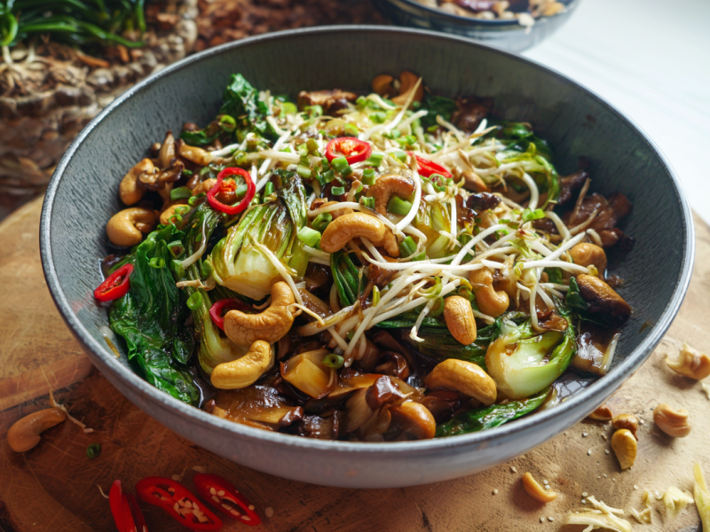 Ein Schüssel mit einer Pak Choi Pilzpfanne auf einem Holzbrett. Um die Schüssel liegen einige Zutaten wie Chilischoten und Cashewkerne verteilt.