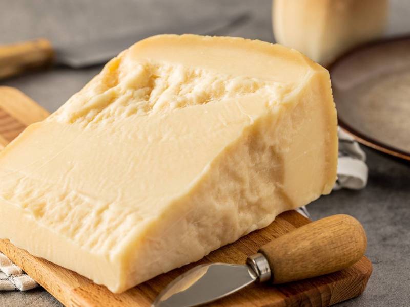 Ein Stück Hartkäse, das auf einem Holzbrett liegt. Ein Käsemesser liegt dabei. Im Hintergrund kann man einen Teller erkennen.