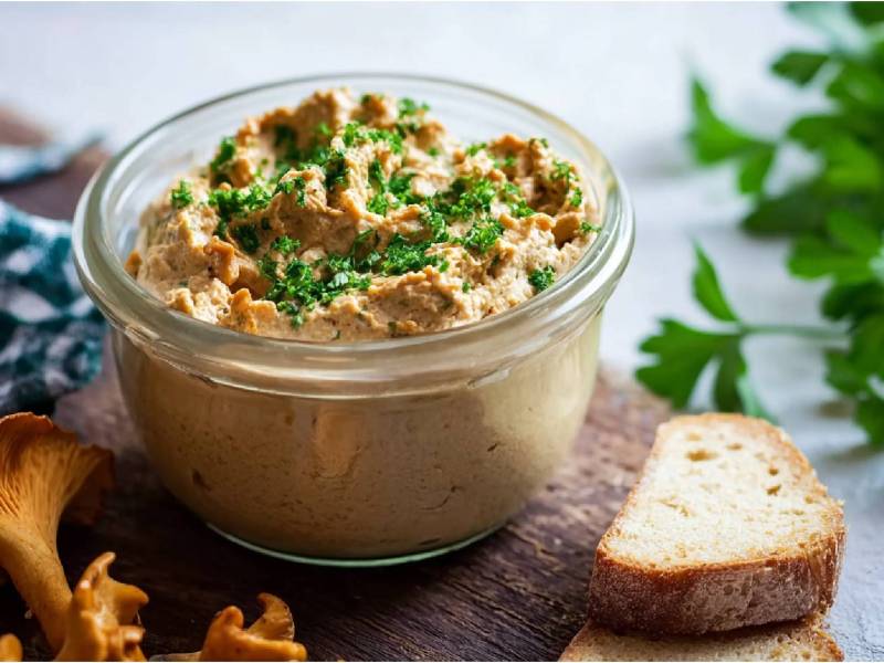 Glas mit Pfifferling-Frischkäse-Aufstrich auf einem Tisch, frische Pfifferlinge und eine Scheibe Brot daneben
