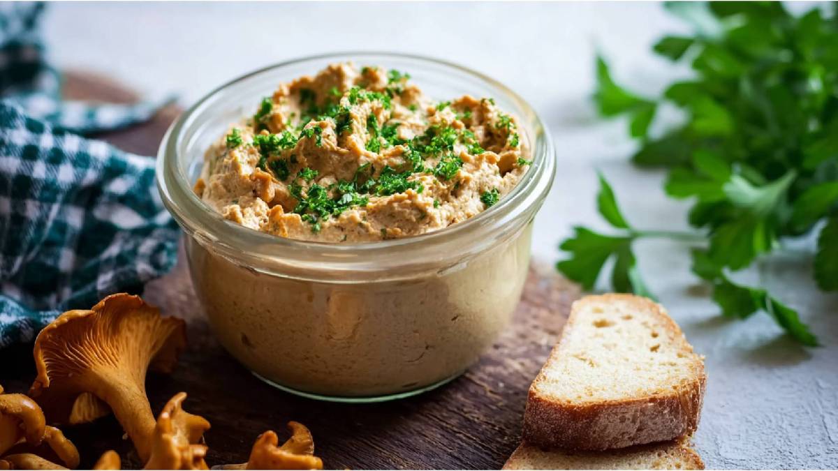 Glas mit Pfifferling-Frischkäse-Aufstrich auf einem Tisch, frische Pfifferlinge und eine Scheibe Brot daneben