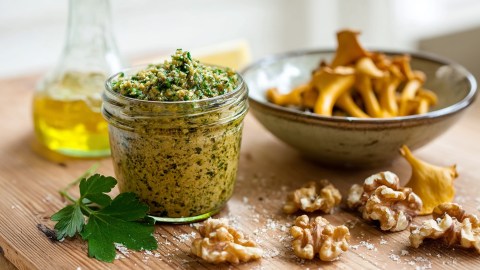 Ein Glas mit Pfifferling-Pesto, dass auf einem Holztisch steht. Im Hintergrund ist eine Schale mit Pfifferlingen und ein Gefäß mit Olivenöl. Außerdem liegt etwas Petersilie und einige Walnüsse herum.
