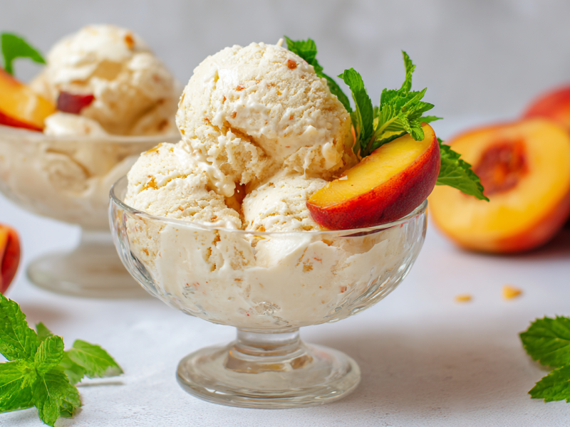 Eine Glasschale mit einem Pfirsich-Joghurt-Eis. Im Hintergrund steht eine weitere Schale sowie mehrere Pfirsiche. Links und rechts sowie in der Schale ist das Eis mit Minzblättern dekoriert.