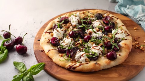 Eine Pizza mit Kirschen und Feta, die auf einem runden Holzbrett liegt. Neben dem Brett liegen einige frische Basilikumblätter und frische Kirschen.