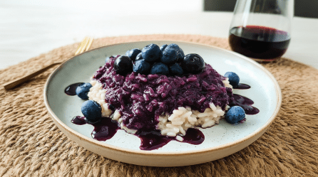 Ein Teller mit Risotto mit Heidelbeeren. Daneben liegt eine Gabel, dahinter steht ein Glas Rotwein.