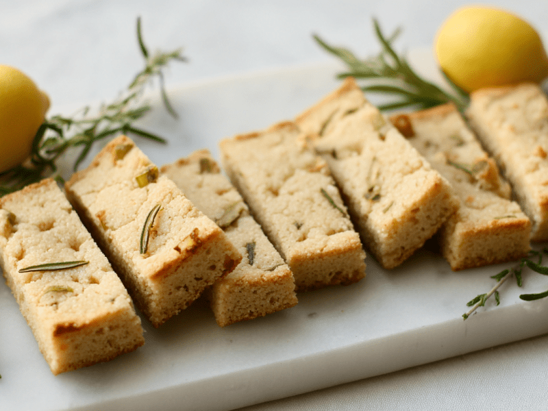 Rosmarin-Zitronen-Shortbread auf einem Marmorbrett, umgeben von Rosmarinzweigen und Zitronen.