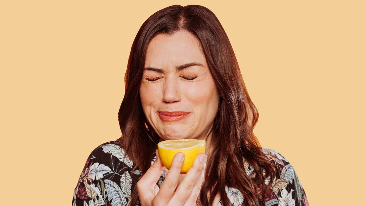 Eine Frau, die eine Grimasse mit geschlossenen Augen zieht, weil sie gerade etwas von einer Zitrone gekostet hat. Die Zitronenhälfte hält sie vor sich. Der Hintergrund ist hellorange.