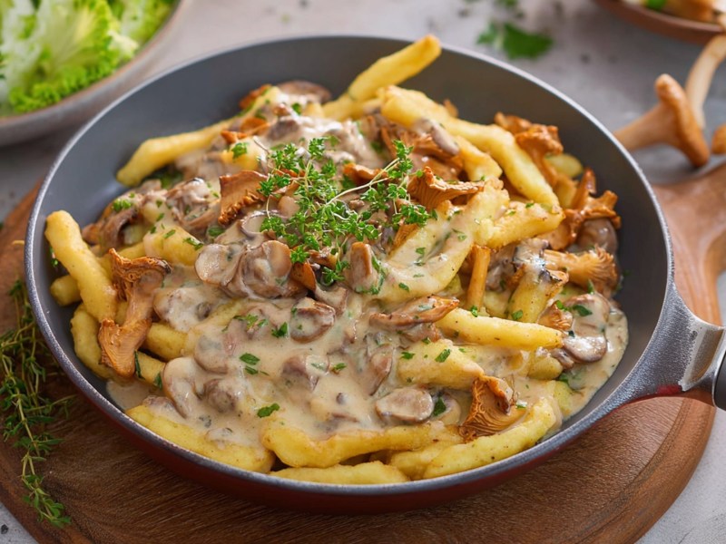 Eine Pfanne mit Schupfnudeln mit Pfifferling-Rahmsoße, die auf einem runden Holzbrett steht. Im Hintergrund kann man einige frische Pfifferlinge erahnen sowie eine Schüssel Beilagensalat.