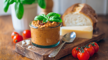 Ein Glas Tomaten-Parmesan-Aufstrich auf einem Holzbrettchen. Im Hintergrund erkennt man Tomaten, Parmesan und Weißbrot.