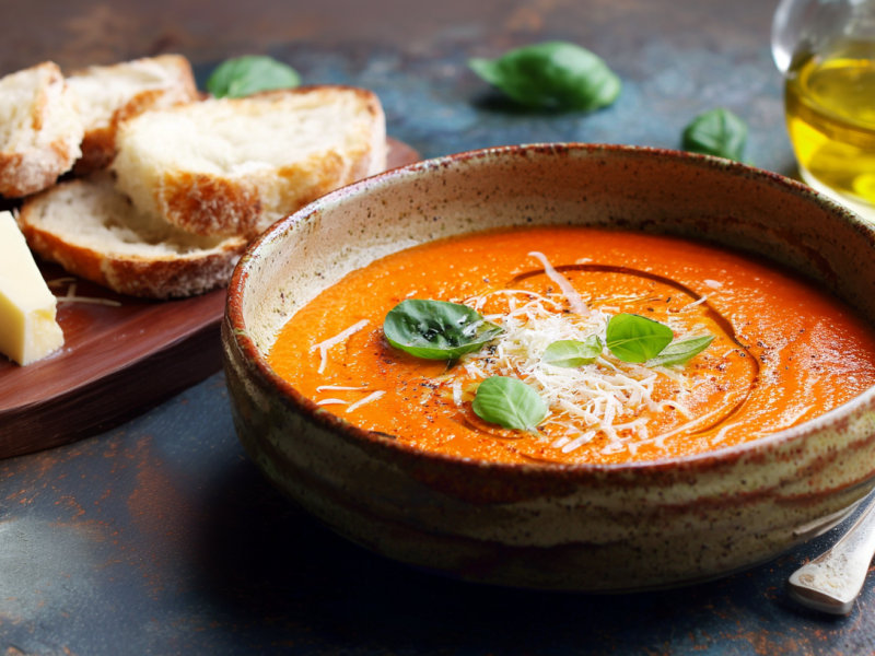 Ein Teller mit Tomaten-Parmesan-Basilikum-Suppe. Daneben steht ein Brett mit Brot und Parmesan, ein kleines Glas mit Olivenöl und ein Löffel liegt daneben.