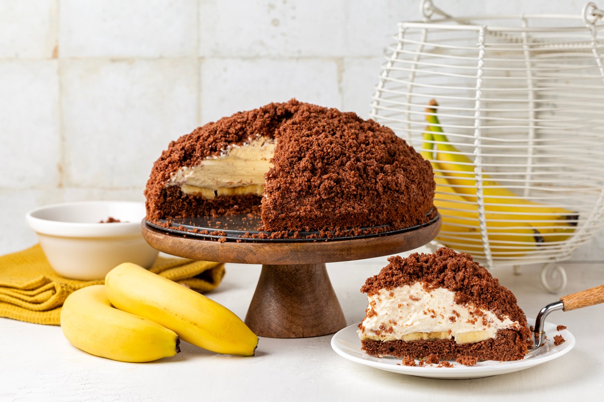Ein veganer Maulwurfkuchen auf einem Präsentierteller. Ein Stück ist herausgeschnitten und gibt den Blick auf die Füllung frei. Davor und dahinter liegen insgesamt vier Bananen.