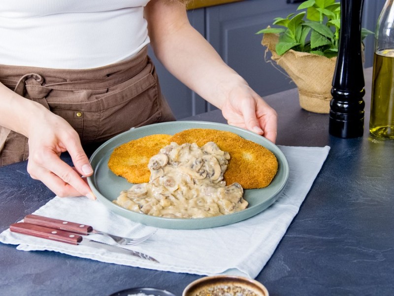 Zwei vegane Jägerschnitzel auf einem grauen Teller. Sie sind mit cremiger Pilzsoße serviert und werden auf einem weißen Tuch präsentiert. Vor dem Teller liegt eine Gabel und ein Messer. Im Hintergrund sieht man frische Kräuter, eine große Pfeffermühle sowie eine Flasche Öl.
