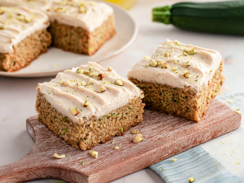 Zwei Zimt-Zucchiniriegel auf einem kleinen Holzbrett. Im Hintergrund sind auf einem Teller noch weitere Riegel. Rechts liegt eine Zucchini.