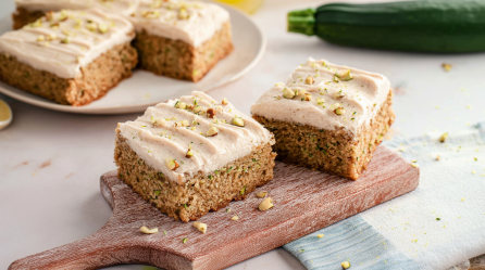 Zwei Zimt-Zucchiniriegel auf einem kleinen Holzbrett. Im Hintergrund sind auf einem Teller noch weitere Riegel. Rechts liegt eine Zucchini.