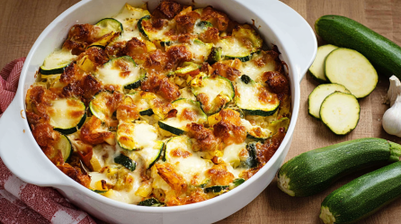 Eine Auflaufform mit Zucchini-Mozzarella-Auflauf, daneben liegen Zucchini.