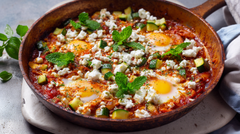 Eine Zucchini-Shakshuka steht auf einem steinernen Untersetzer. SIe ist mit frischer Minze garniert. Links neben der Pfanne liegt etwas Grünzeug, rechts ein Küchentuch.