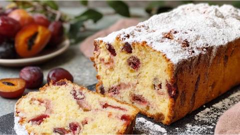 ein angeschnittener Zwetschgen-Joghurt-Kuchen aus der Kastenform auf einem Tisch, Zwetschgen daneben