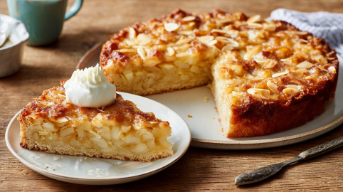 Ein Stück Apfel-Bienenstich auf einem kleinen Teller mit etwas Sahne darauf. im Hintergrund sieht man den restlichen Kuchen.