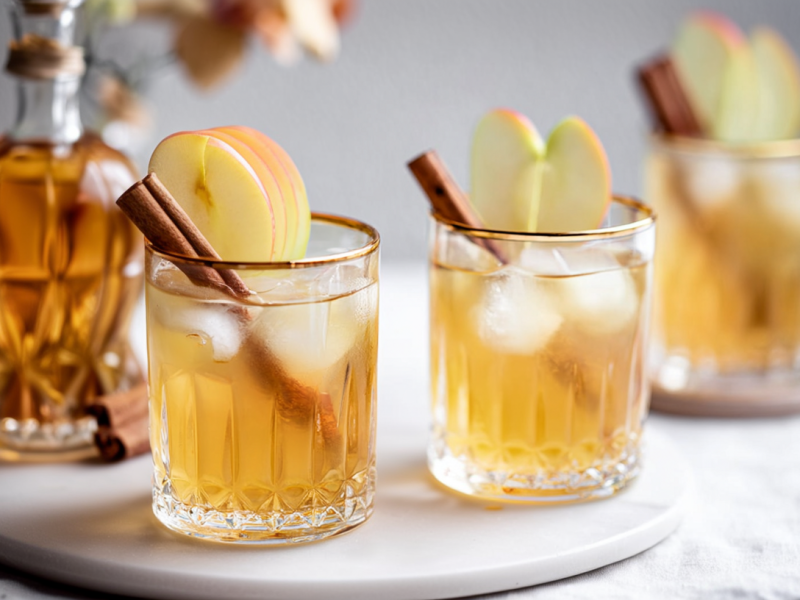 Zwei Gläser mit Apfel Lillet stehen auf einem Tablett. Garniert sind die Drinks mit Zimtstangen und Apfelscheiben. Im Hintergrund ist ein weiteres Glas sowie eine Karaffe zu sehen.