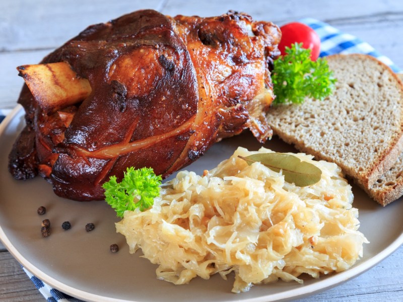 Eine bayerische Schweinshaxe mit Sauerkraut und Brotscheiben auf einem Teller.