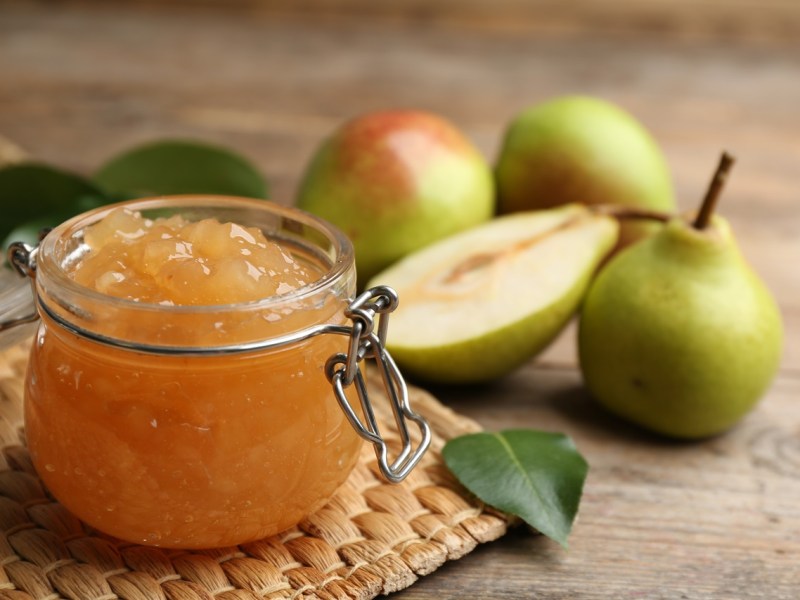 Ein Glas mit Birnen-Marzipan-Marmelade steht auf einem Platzdeckchen. Im Hintergrund sind Birnen zu sehen.