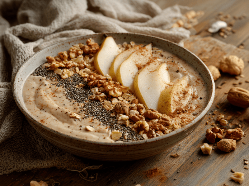 Eine Birnen-Zimt-Smoothie-Bowl auf einem Holztisch. Um sie herum verteilt liegen Walnüsse und etwas Zimt. Sie steht direkt an einem grau-braunen Tuch.