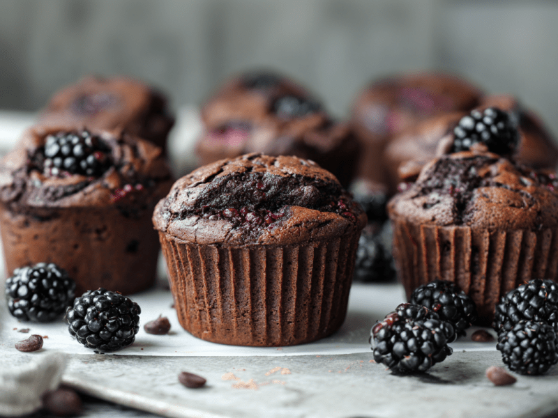 Mehrere Brombeer-Schoko-Muffins sind auf einem Tablett angerichtet. Frische Beeren liegen als Deko außen herum.